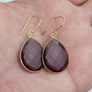 May include: A pair of gold dangle earrings with teardrop-shaped amethyst gemstones. The gemstones are faceted and have a deep purple hue.