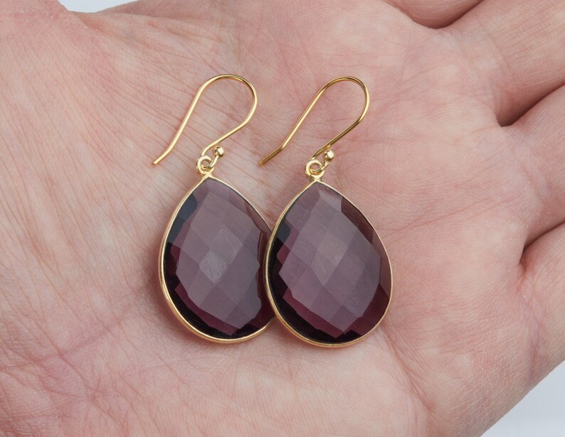 May include: A pair of gold dangle earrings with teardrop-shaped amethyst gemstones. The gemstones are faceted and have a deep purple hue.