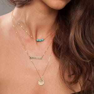 May include: A layered gold necklace set. The top layer features small gold beads and turquoise stones. The middle layer has a rectangular bar pendant. The bottom layer has a textured, circular pendant. The necklaces are worn on a person.