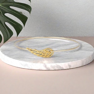 May include: A gold bangle bracelet with a delicate wing design. The bracelet is on a white marble surface.