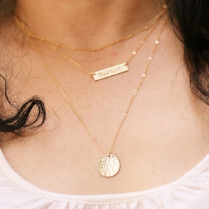 Puede incluir: Un collar de oro de varias capas con un pequeño colgante de disco de oro martillado y un colgante de barra de oro más largo y delgado con el texto "10.27.114" grabado en él.