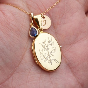 May include: Gold locket necklace with a delicate floral design and a small blue gemstone charm. The locket has a personalized initial charm.