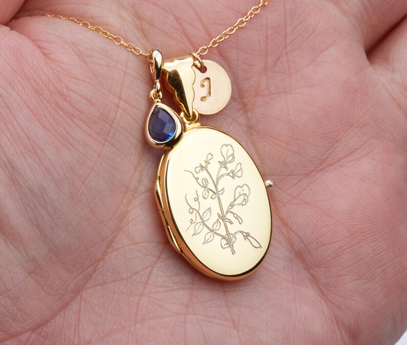 May include: Gold locket necklace with a delicate floral design and a small blue gemstone charm. The locket has a personalized initial charm.