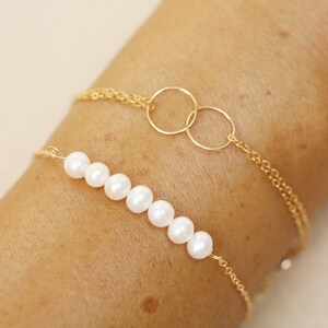 Könnte beinhalten: Ein goldenes Kettenarmband mit zwei ineinander verschlungenen Kreisen und einer Reihe weißer Perlen.