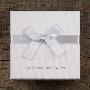 May include: A white gift box with a silver ribbon and bow. The box has the text "Tyrahandmadejewelry" printed on the front.