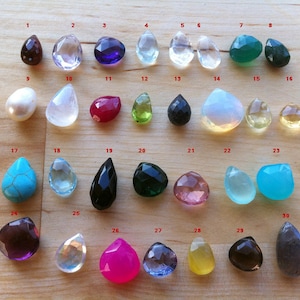 Puede incluir: Treinta colgantes de piedra preciosa en forma de l&aacute;grima de diferentes colores, incluyendo morado, verde, azul, rosa y blanco. Los colgantes est&aacute;n dispuestos en filas sobre una superficie de madera.