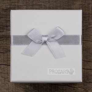 Puede incluir: Una caja de regalo blanca con una cinta y un lazo plateados. La caja tiene la palabra "PROZART" impresa.
