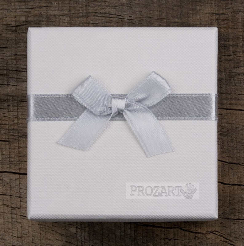 Puede incluir: Una caja de regalo blanca con una cinta y un lazo plateados. La caja tiene la palabra "PROZART" impresa.