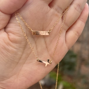 Puede incluir: Collar de oro con un delicado colgante de cruz y un colgante de barra que dice "HOPE".
