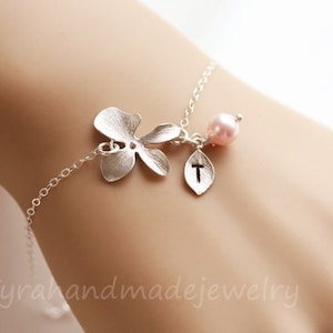 May include: A silver bracelet featuring a delicate orchid charm, a pink pearl, and a leaf charm engraved with the letter "T".