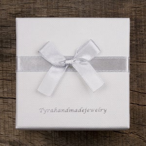 Może przedstawiać: Białe pudełko prezentowe z srebrną wstążką i kokardką. Na przodzie pudełka znajduje się napis "Tyrahandmadejewelry".