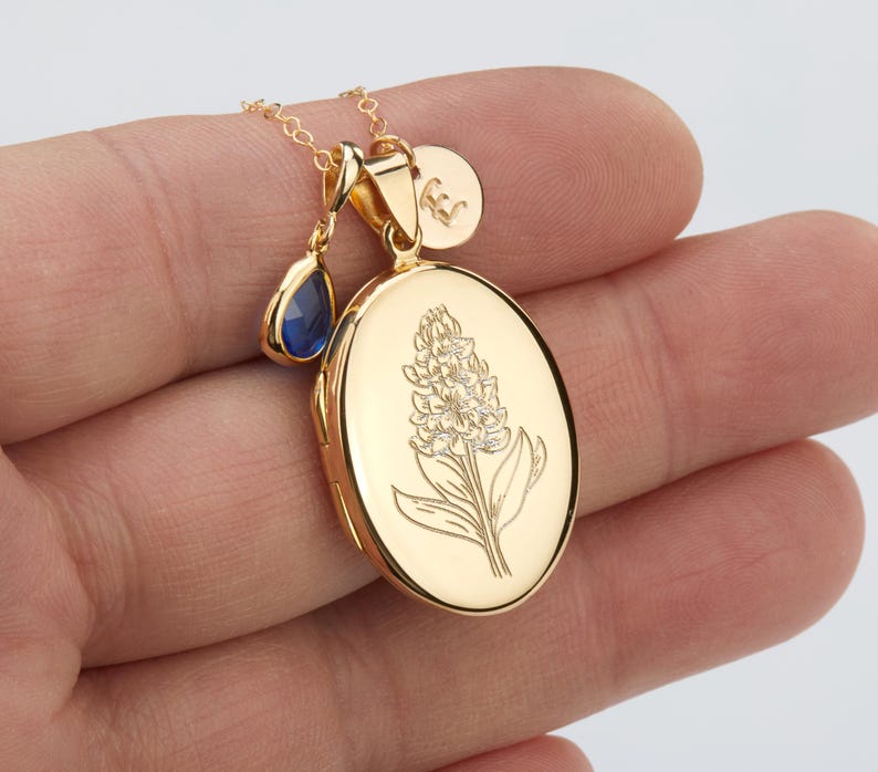 May include: A gold locket necklace with a floral design and a blue teardrop charm. The locket is oval shaped and has a delicate chain.