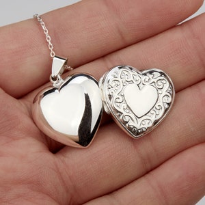 May include: Silver heart-shaped locket necklace with a detailed filigree design on the front. The locket opens to reveal a smooth, blank interior.