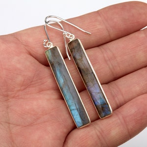 Puede incluir: Un par de pendientes de plata con piedras rectangulares de labradorita largas. Las piedras tienen una base gris oscuro con destellos iridiscentes azules y verdes.