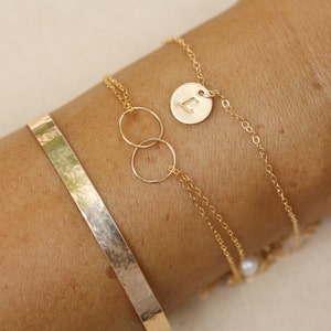 Peut inclure: Un ensemble de bracelets en or comprenant une chaîne délicate avec un petit pendentif rond, un pendentif double cercle et un bracelet manchette martelé. Le bracelet à chaîne a un petit charme avec la lettre "L" dessus.