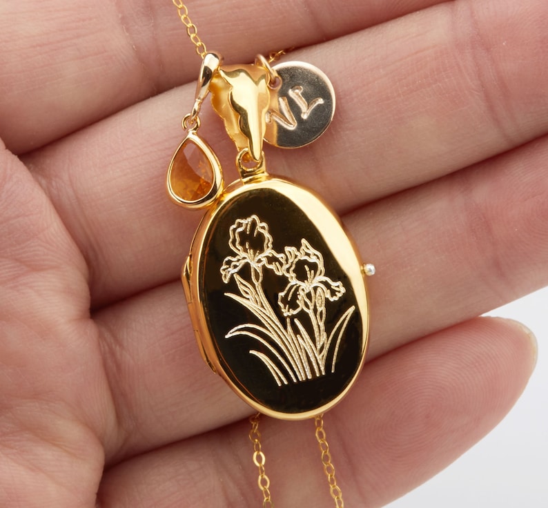 May include: Gold locket necklace with a delicate chain, featuring a small teardrop-shaped gemstone and an engraved oval locket with a floral design. The locket has a small charm with the letter "N" on it.