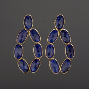 May include: A pair of gold-toned earrings featuring a cluster of oval-shaped, faceted, deep blue gemstones. The gemstones are arranged in an open, elongated oval design. The earrings are set against a dark gray background.