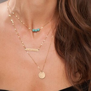 May include: A layered gold necklace set. The top layer features three turquoise beads. The middle layer has a rectangular bar with engraved text. The bottom layer has a round pendant. The necklace is worn on a person.