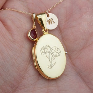 May include: Gold locket necklace with a small red gemstone charm and a personalized initial charm. The locket features an engraved floral design.