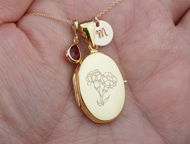 May include: Gold locket necklace with a small red gemstone charm and a personalized initial charm. The locket features an engraved floral design.