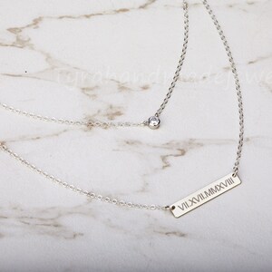 May include: A silver chain necklace with a small clear stone charm and a bar pendant with the Roman numeral inscription "VII.XVII.MMXVIII".