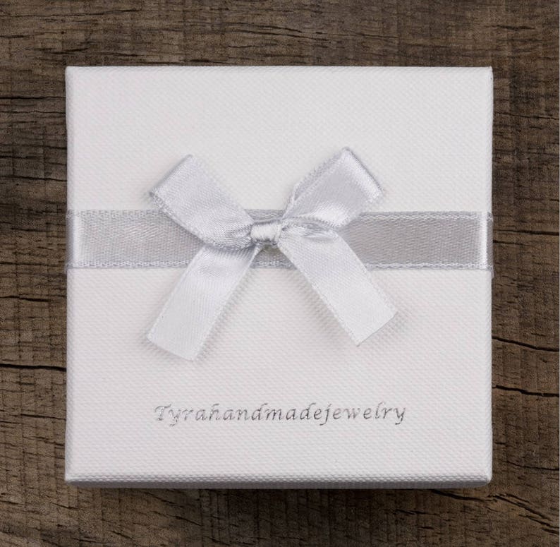 May include: A white jewelry box with a textured surface, adorned with a silver ribbon and bow. The text "Tyrahandmadejewelry" is printed on the box. The box is set against a wooden background.