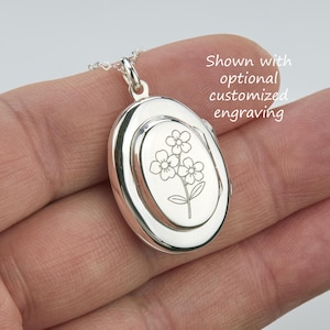 Può includere: Un ciondolo a medaglione ovale in argento lucido su una catenina delicata, caratterizzato da un'incisione floreale centrale di tre piccoli fiori con foglie. Il testo "Shown with optional customized engraving" è visibile in alto a destra.