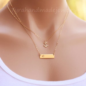 May include: A gold layered necklace with a small anchor charm and a bar pendant with the letter 'C' engraved on it.