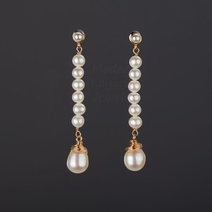 Puede incluir: Un par de pendientes largos de perlas blancas con detalles de alambre dorado. Las perlas están enhebradas en una cadena dorada y la perla inferior es más grande que las demás.