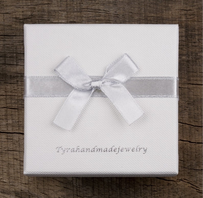 May include: A white gift box with a silver ribbon and bow. The box has the text "Tyrahandmadejewelry" printed on the front.