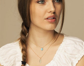 May include: A woman wearing a white shirt with a gold chain necklace featuring a turquoise teardrop pendant and a gold leaf pendant.