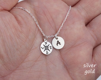 May include: A silver necklace with a compass charm and a charm with the letter A. The necklace is on a hand.