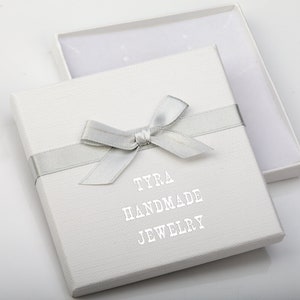 May include: White gift box with a silver ribbon and bow. The box has the text "TYRA HANDMADE JEWELRY" printed on the front.