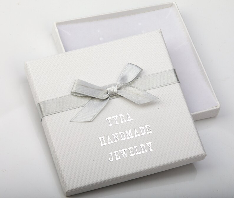 May include: White gift box with a silver ribbon and bow. The box has the text "TYRA HANDMADE JEWELRY" printed on the front.