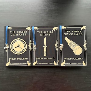 Peut inclure: Trois livres reliés de la série "À la croisée des mondes", avec des titres dorés et des illustrations sur des couvertures noires. Chaque livre a un signet en ruban bleu avec un charme. Titres : "La boussole d'or", "La tour des anges", et "Le miroir d'ambre".