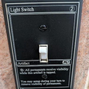 May include: Black light switch cover with the text "Light Switch" and "Artifact". The switch has a white toggle and is mounted on a wall. The text "All permanents receive visibility while this artifact is tapped" is also visible.