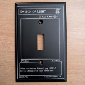 May include: A matte black rectangular light switch cover plate designed to resemble a trading card. It features white printed text including "SWITCH OF LIGHT," "TRAP CARD," "1st Edition," "LZR-023," and game instructions with two screw holes.