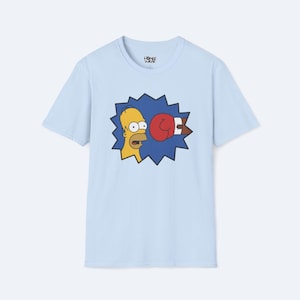 May include: Light blue t-shirt featuring a cartoon image of Homer Simpson being punched by a red boxing glove. The graphic is set against a blue burst background. The shirt has a crew neck and short sleeves.