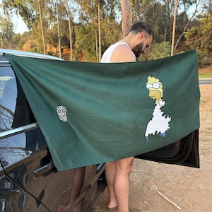 May include: A dark green beach towel featuring a cartoon image of Homer Simpson with a white background. The towel has Japanese text in the upper left corner and is being held up near a black car.