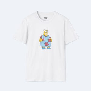 May include: White t-shirt featuring a cartoon illustration of Homer Simpson in a Hawaiian shirt and a white hat. The shirt has a crew neck and short sleeves. The Homer Simpson character is wearing a blue shirt with pink flowers.