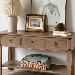 Può includere: Un tavolo console in legno chiaro con tre cassetti e uno scaffale inferiore. Il tavolo ha una lampada con un paralume color crema, un quadro incorniciato e una pila di libri in cima. Un cesto di vimini con un panno verde all'interno si trova sullo scaffale inferiore.
