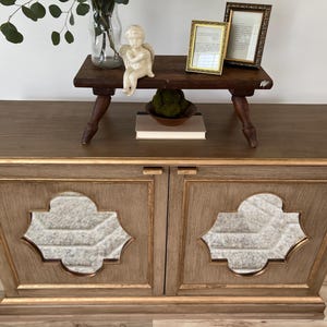 May include: A wooden cabinet with two doors, each featuring a decorative, gold-trimmed, quatrefoil-shaped mirror. The cabinet has a light brown finish and gold accents. A small wooden table with a figurine of an angel sits on top of the cabinet.