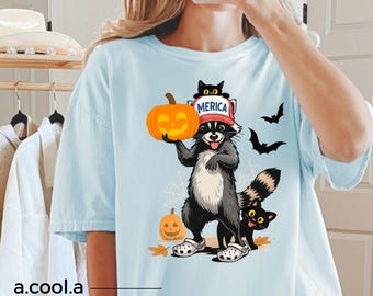Op de afbeelding: Een lichtblauw t-shirt met een afbeelding van een wasbeer die een rood-wit-blauwe pet draagt met de tekst MERICA. De wasbeer houdt een oranje pompoen vast, heeft een zwarte kat op zijn hoofd en een andere zwarte kat bij zijn voeten.