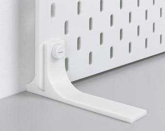 May include: A white pegboard with evenly spaced, vertical oval holes. A white bracket supports the board, attached with a circular fastener. The bracket extends horizontally, resting on a gray surface. The pegboard is designed for organization and display.