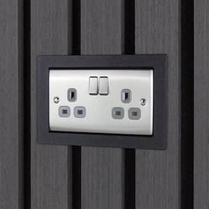Op de afbeelding: Een dubbel elektrisch wandstopcontact met een metallic zilveren afwerking en twee schakelaars, gemonteerd in een zwart rechthoekig frame op een donkergrijze wandplaat met verticale latten.