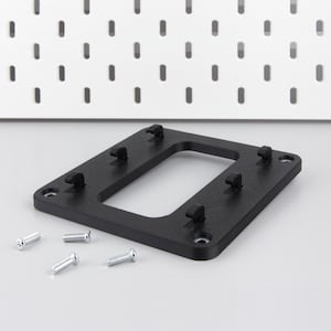 Op de afbeelding: Een zwarte montagebeugel voor pegboard met vier verhoogde lipjes en vier zilveren schroeven. De beugel heeft een rechthoekige uitsparing. Een wit pegboard met gelijkmatig verdeelde gaten staat op de achtergrond.