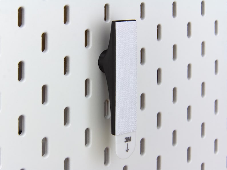 Pu&ograve; includere: Un gancio nero per pannello forato con una striscia adesiva bianca, progettato per il montaggio su un pannello forato bianco. Il gancio &egrave; in plastica nera e ha un design curvo. La striscia adesiva presenta il logo "3M" e una freccia che indica la direzione di applicazione.