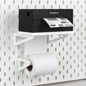 May include: A black MUNBYN thermal label printer sits on a white wall-mounted shelf. A roll of white thermal labels is held by a white bracket underneath the shelf, with a printed shipping label feeding into the printer from the roll below.