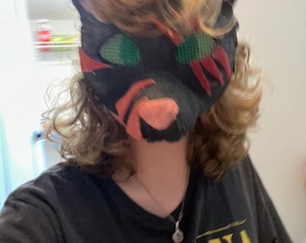 May include: A black cat mask with red, green, and pink accents. The mask is worn over a black t-shirt with yellow text that reads "NIRVANA". The mask has large, pointed ears and is made of a soft material.