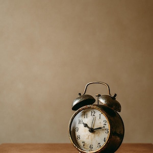 May include: Vintage alarm clock with a distressed, aged appearance. The clock features a round face with black hands and numerals, set on a brown wooden surface. The clock's metal casing has a tarnished, antique finish, with two bells on top.
